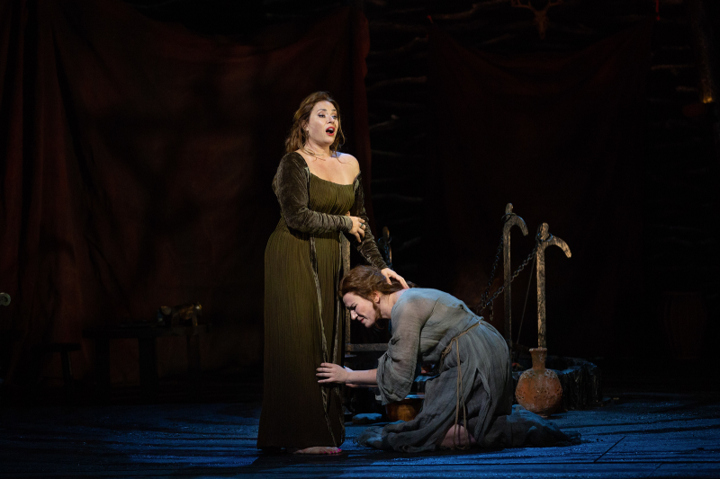   Una mujer angustiada se arrodilla ante una mujer que canta en una representación operística.