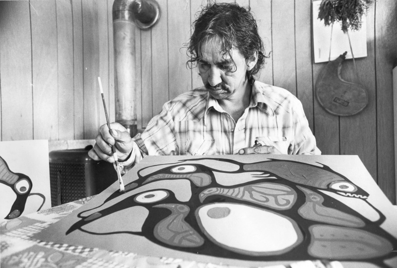  Imagen en blanco y negro de Norval Morrisseau sentado en un escritorio, pintando un lienzo frente a él